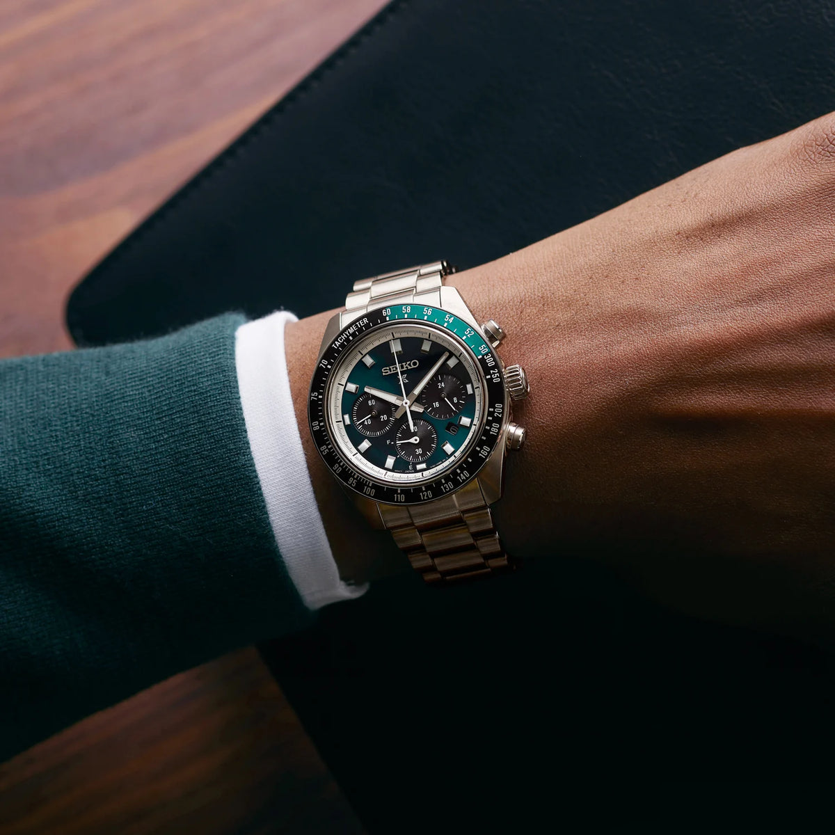 A wrist wearing a stainless steel chronograph watch with a black dial, silver bezel, and a distinctive green accent ring around the bezel. The watch is shown on a person's wrist wearing a dark green sweater over a white collared shirt.