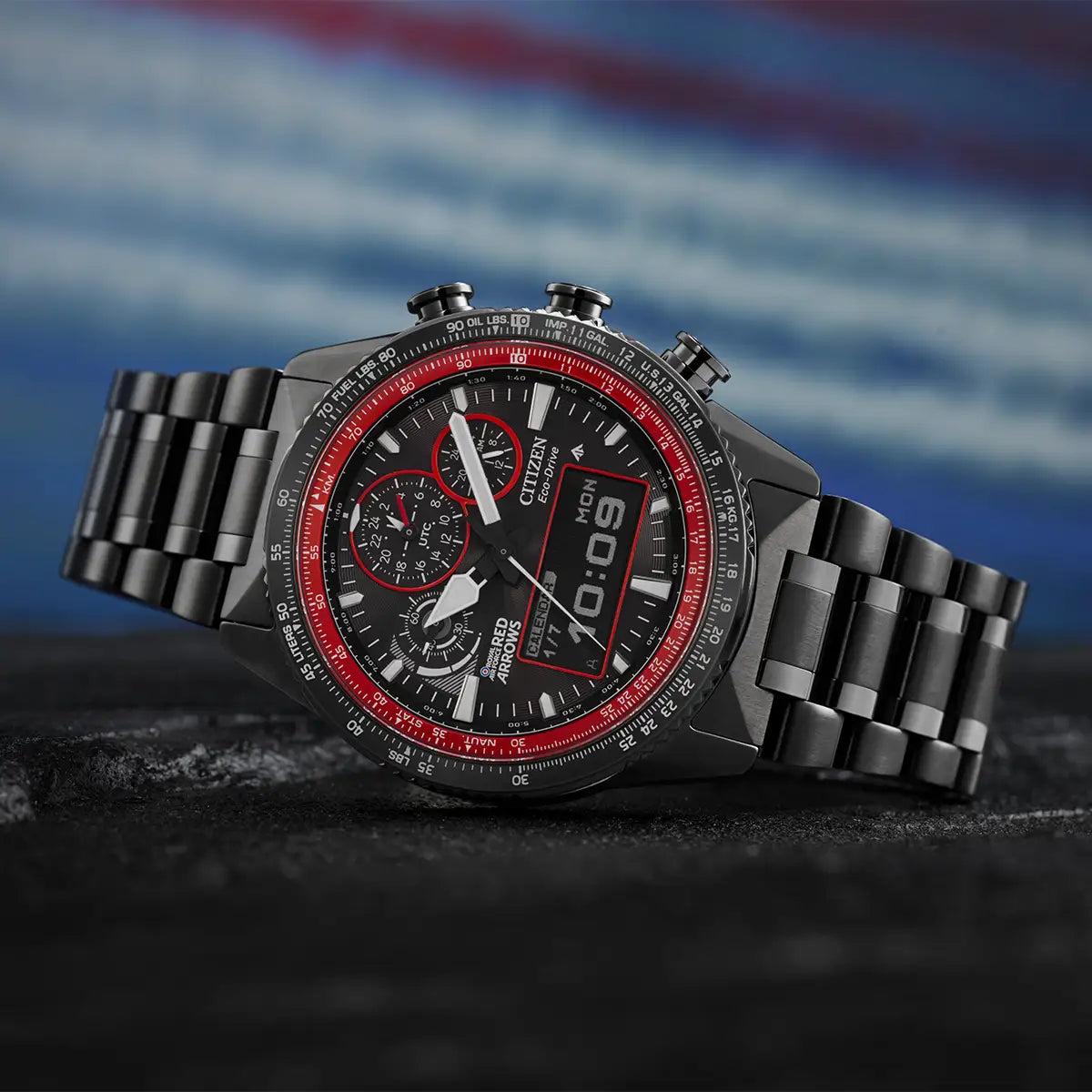 A rugged chronograph watch with a black metal link bracelet, black dial, and red accents, resting on a dark textured surface with a blurred blue background.
