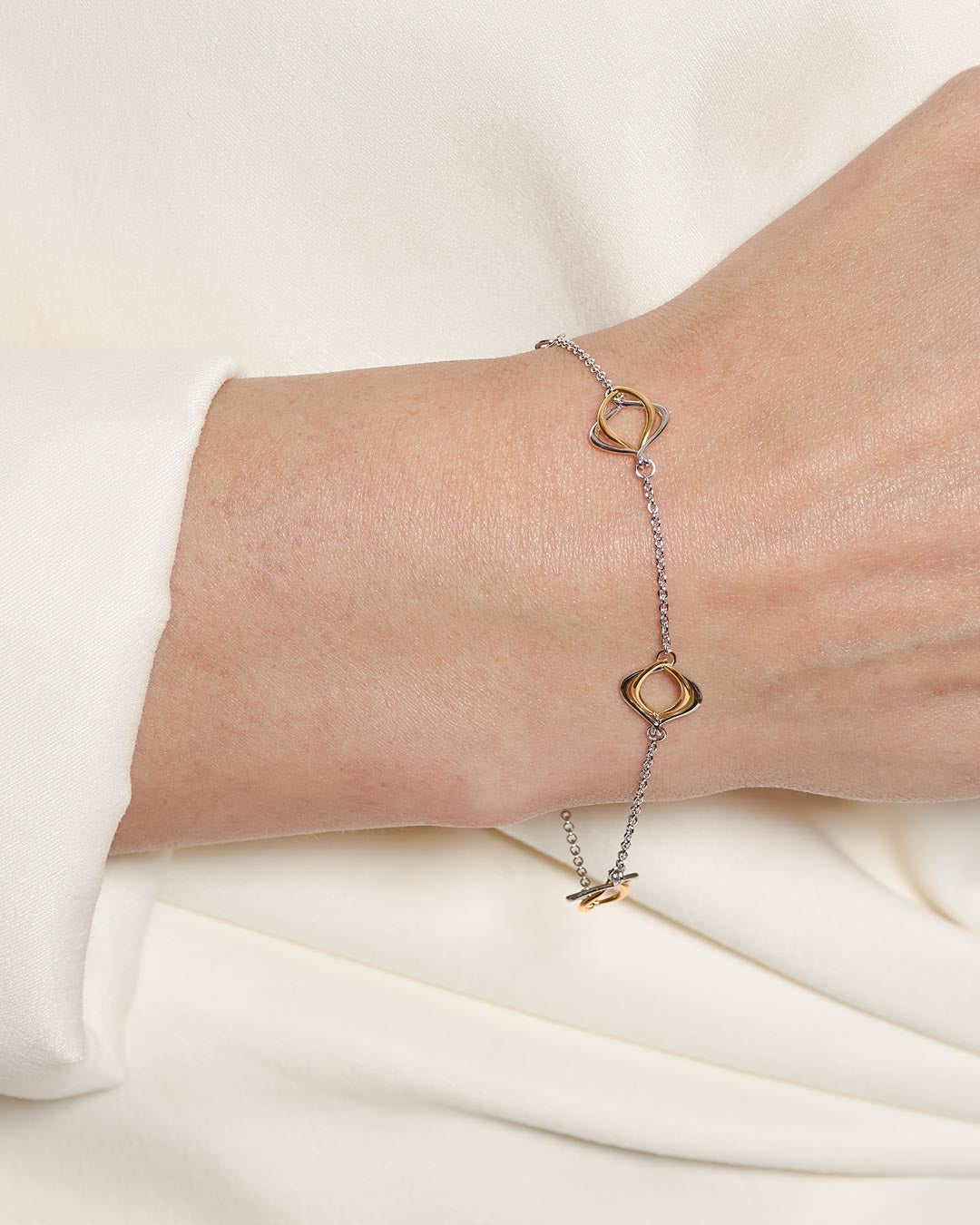 A delicate two-tone chain bracelet with gold and silver interlocking oval links, shown worn on a person's wrist against a soft cream-colored fabric background.