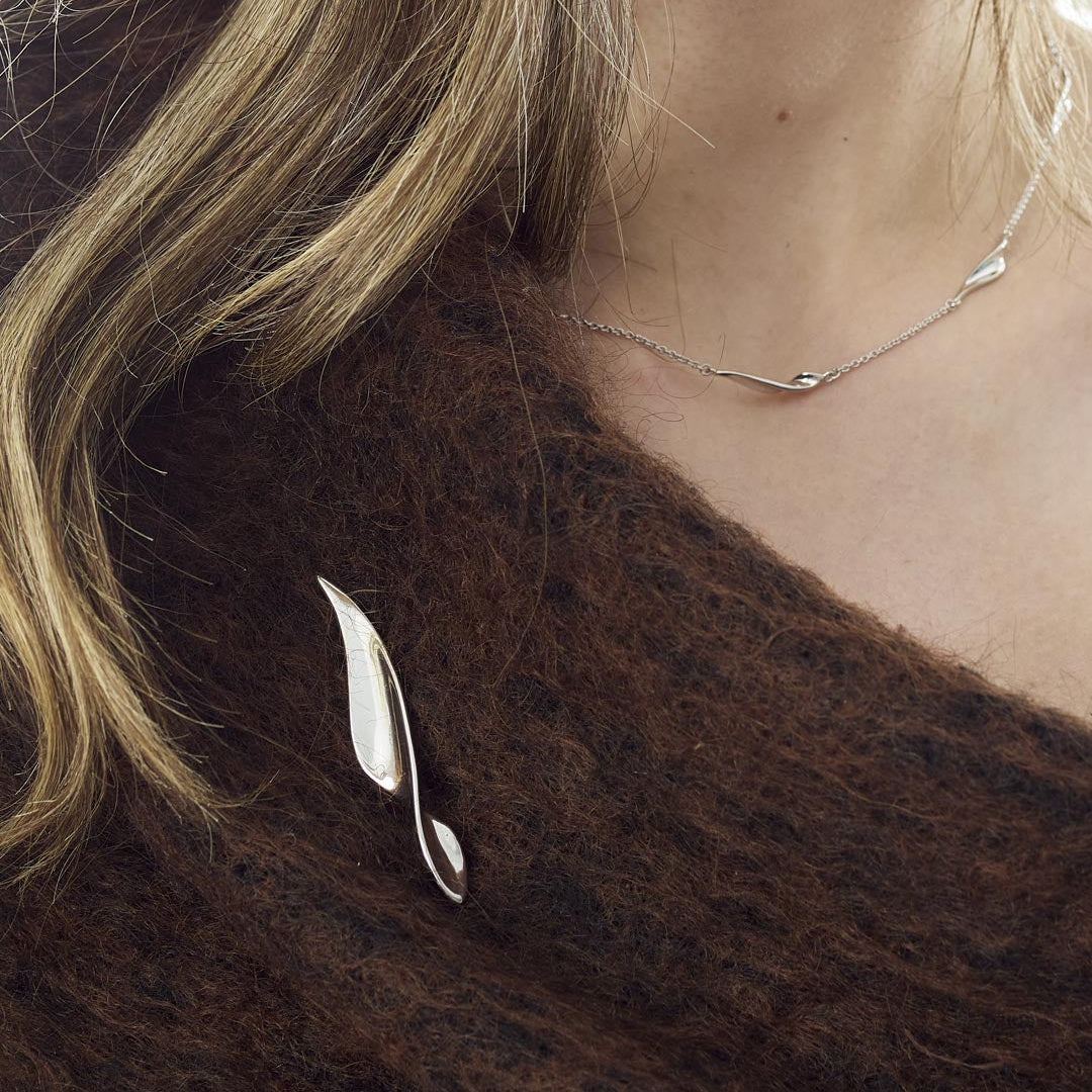 A close-up of a person wearing a silver leaf-shaped brooch on a dark brown textured sweater, with a delicate silver necklace also visible.